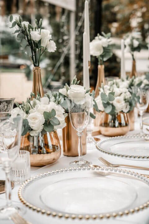 Elegante Hochzeitstischdekoration mit weißen Rosen, grünen Blättern, goldenen Vasen und Kristallgläsern für Champagnerempfang