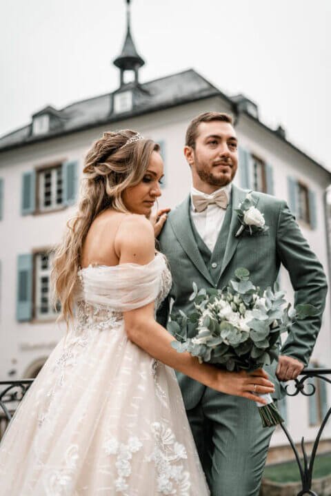 Brautpaar in festlicher Kleidung steht vor historischem Gebäude mit einem Strauß aus Eukalyptus und weißen Blumen