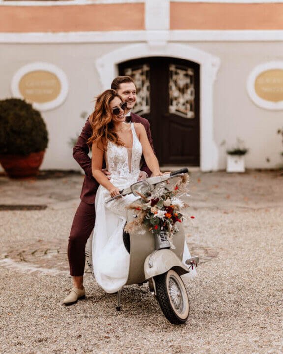 Brautpaar auf einer Vespa vor historischem Gebäude, lachend und festlich gekleidet für eine Hochzeitsfeier