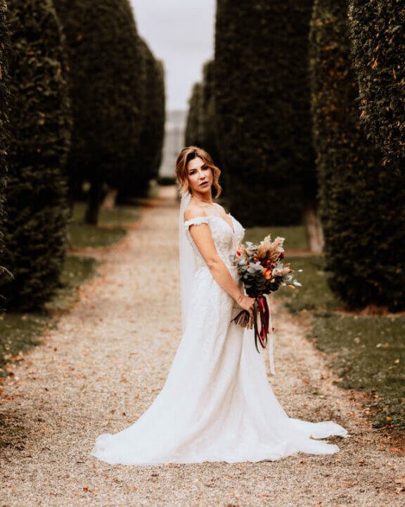 Braut in elegantem Hochzeitskleid mit Blumenstrauß steht auf einem Kiesweg umgeben von hohen Hecken