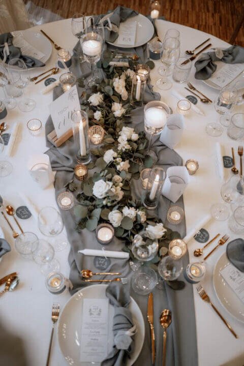 Elegante Hochzeitstafel mit weiß-blumigem Centerpiece, Goldbesteck und grauen Servietten