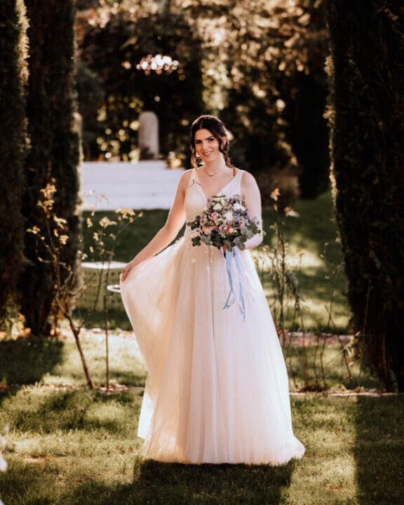 Braut in einem fließenden Hochzeitskleid hält einen bunten Blumenstrauß in einem idyllischen Garten