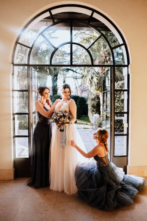 Braut in einem eleganten weißen Kleid mit einem farbenfrohen Blumenstrauß, flankiert von zwei Brautjungfern in festlichen Kleidern während einer stilvollen Hochzeitsvorbereitung in einem sonnendurchfluteten Raum mit Blick auf einen Garten