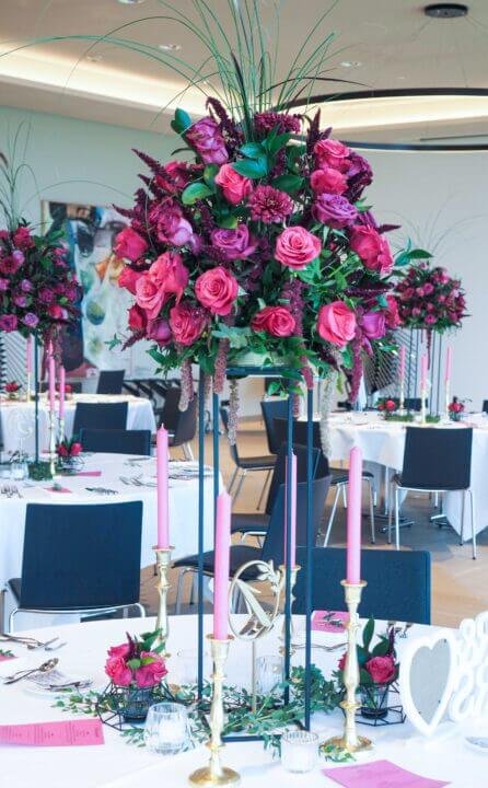 Elegante Hochzeitstischdekoration mit üppigem Blumengesteck aus rosa Rosen und dunkelroten Akzenten, goldenen Kerzenhaltern und feinem Geschirr in einem festlich geschmückten Raum