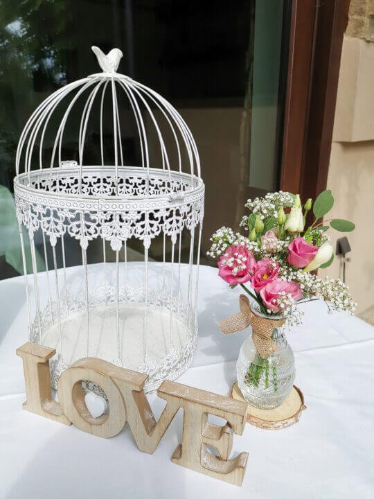 Dekorative Hochzeitstischdekoration mit einem weißen Vogelkäfig, einem Blumenstrauß in einer Vase und einem Holzschriftzug Love