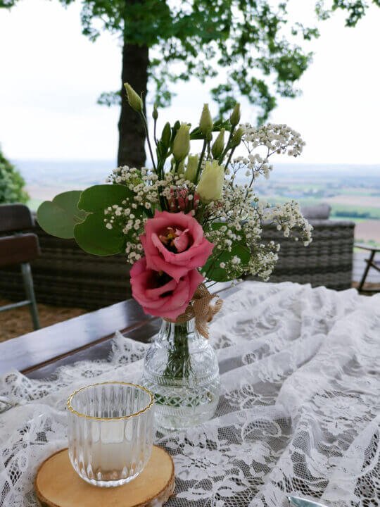 Blumenarrangement mit rosafarbenen Blumen und Spitzentischdecke bei einer Außendekoration für eine Hochzeit